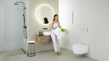 A fürdőszobában mosdókagylóra támaszkodó nő Geberit AquaClean Sela zuhany WC-vel és Geberit VariForm mosdókagylóval és bútorokkal