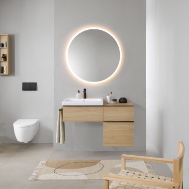 Fürdőszoba szürke falakkal, Geberit fa fürdőszobabútorokkal és egy kerek Geberit Option világító tükörrel.
